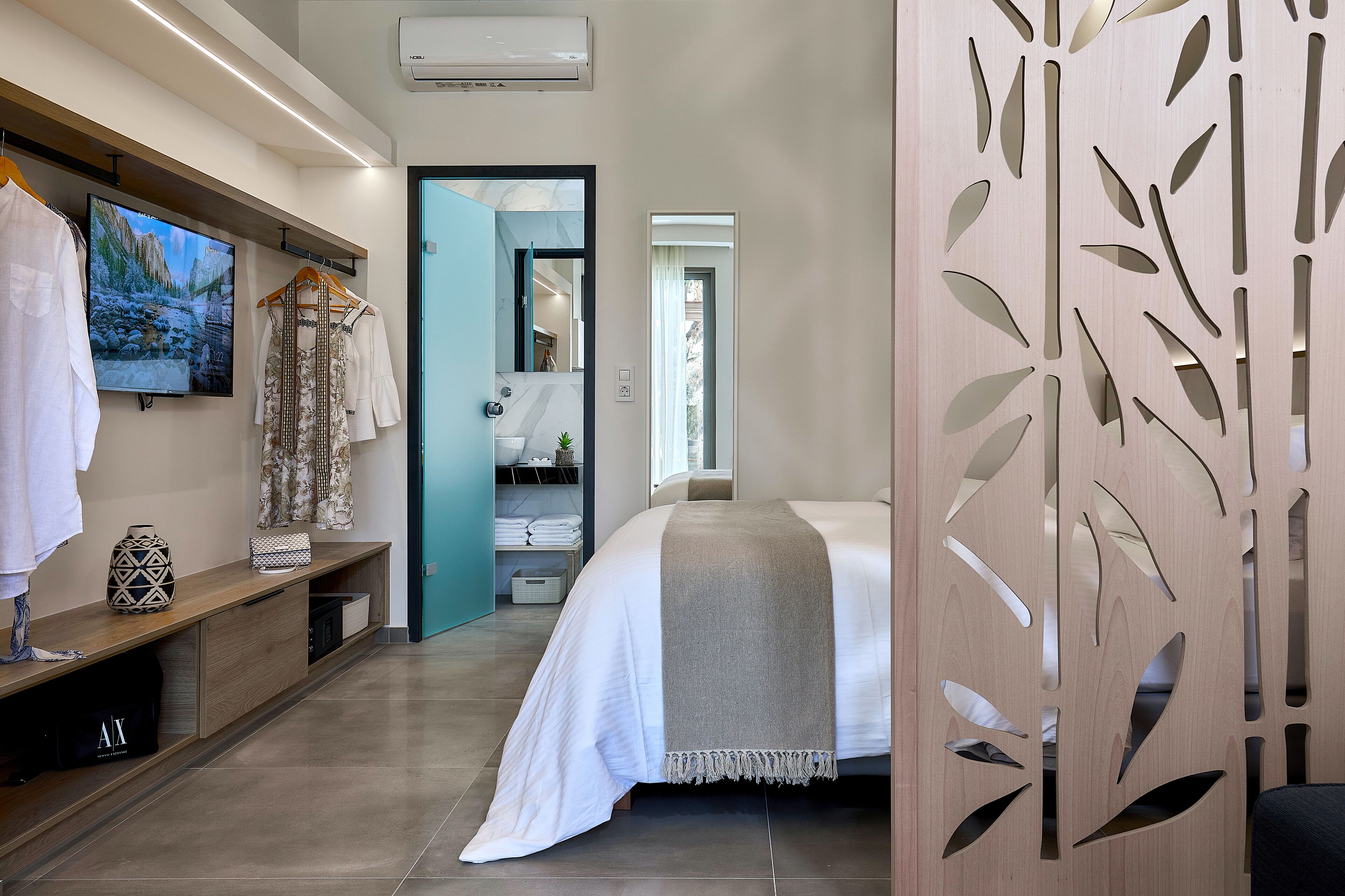 A modern hotel room with a bed, a TV, a wooden bench, and a decorative panel.