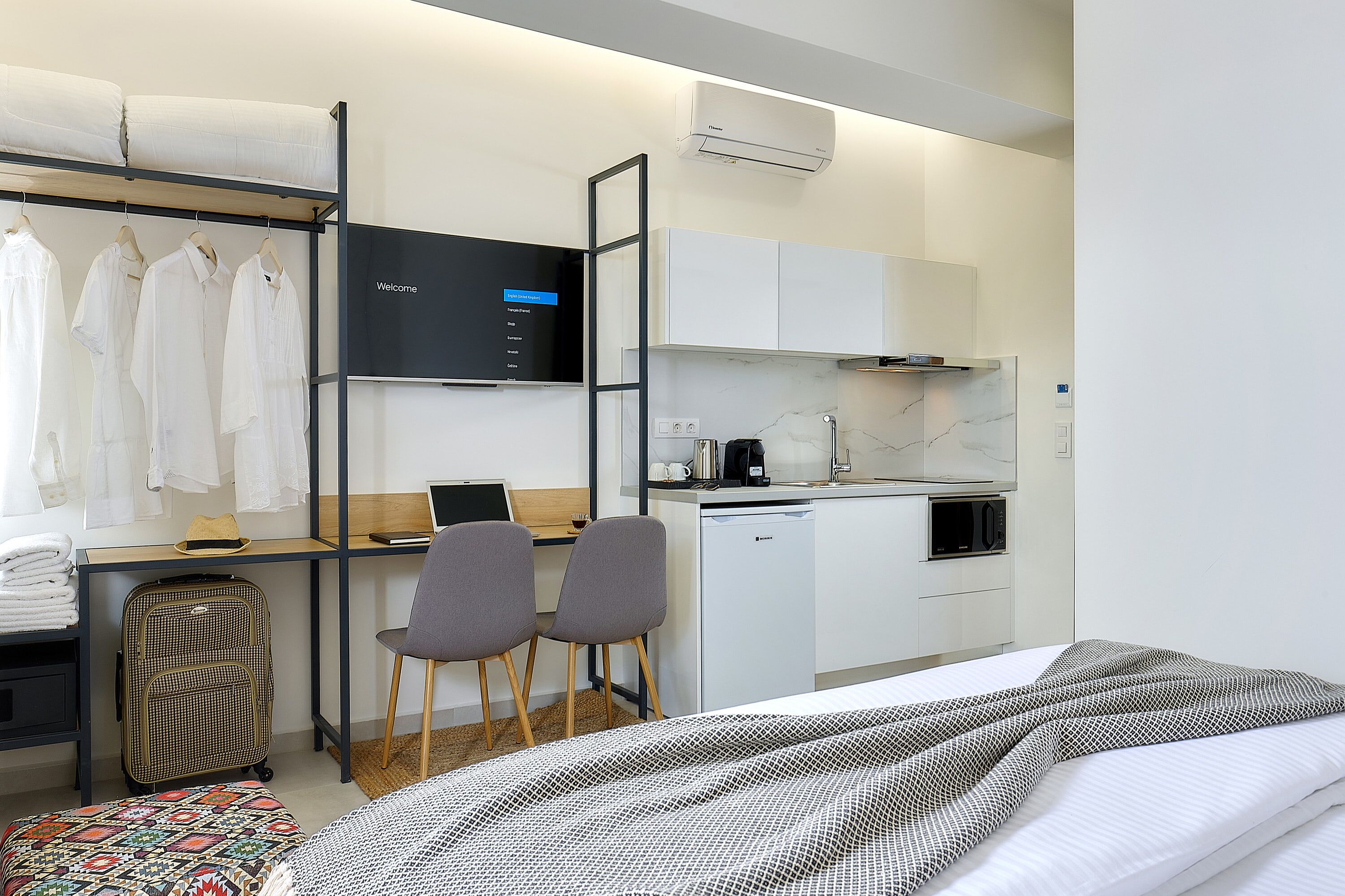 Une chambre d’hôtel moderne équipée d’un lit, d’un bureau avec un ordinateur portable, d’une petite kitchenette et d’une armoire avec des vêtements suspendus.