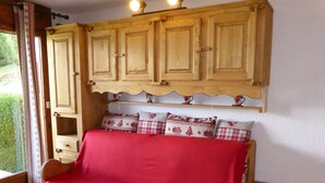 A cozy room with a wooden sofa bed covered in a red blanket, wooden cabinets above, and a small wooden table beside the bed. - Studio for 4, opposite ski lift, private terrace (Combloux)