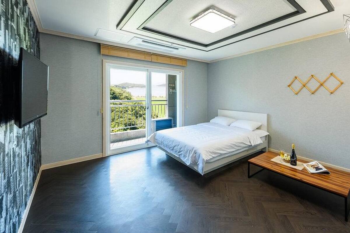 Une chambre à coucher avec un lit, une table de chevet sur laquelle se trouve une bouteille, un téléviseur fixé au mur et un balcon avec vue.