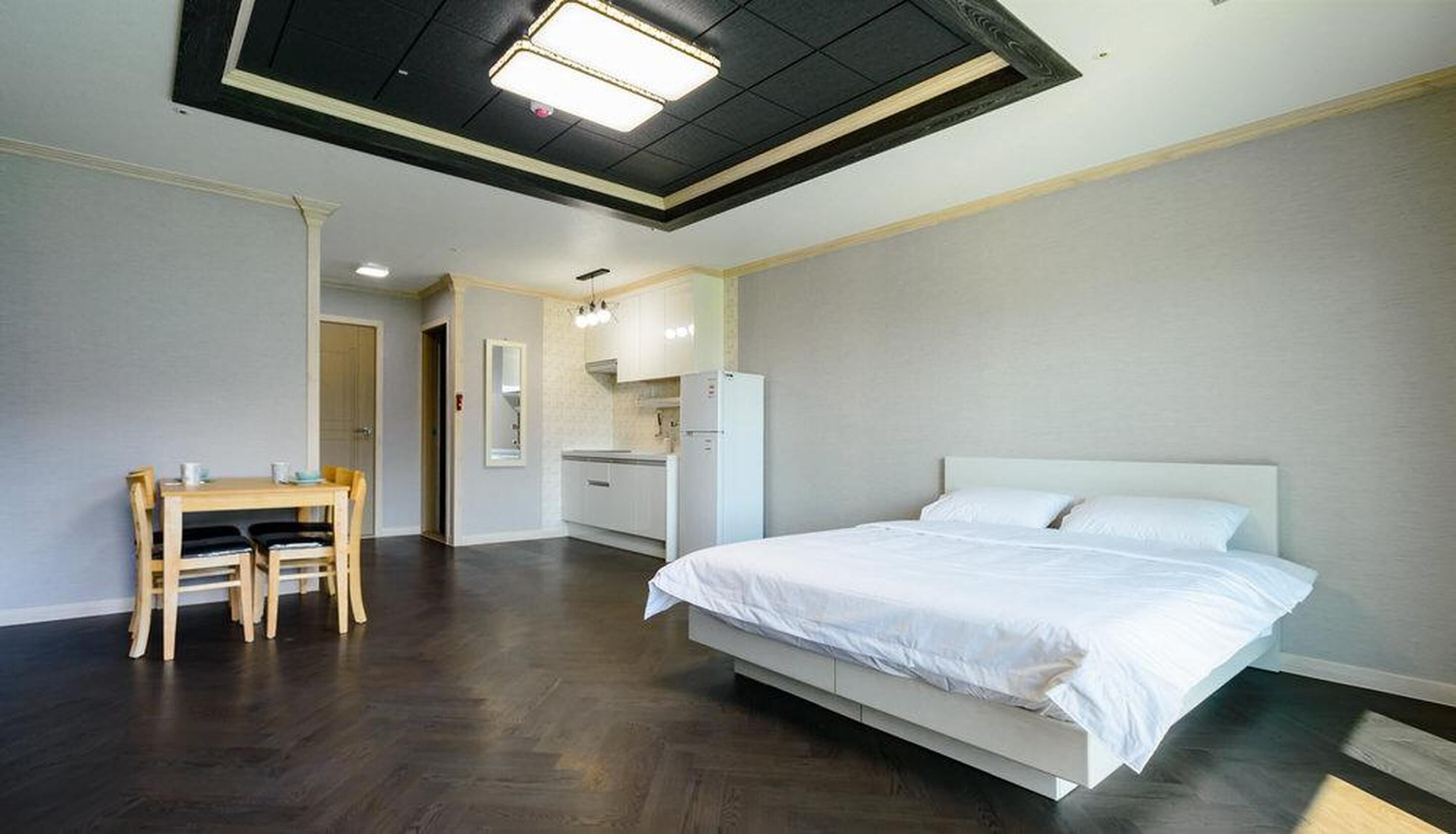 Ett modernt hotellrum med en stor säng, ett matbord med stolar, ett litet kök och ett tak med infälld belysning.
