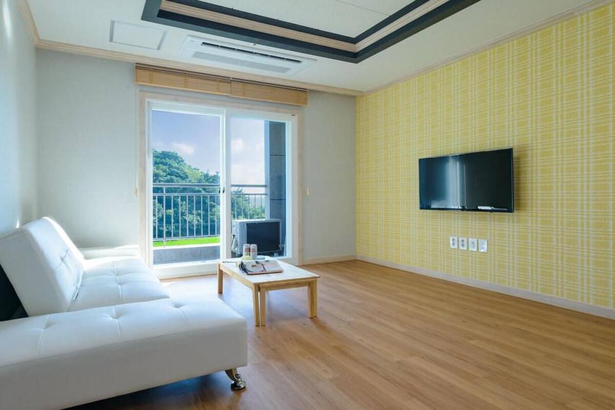 Un salon moderne avec un canapé blanc, une petite table, un téléviseur à écran plat fixé au mur et un balcon donnant sur un paysage verdoyant.