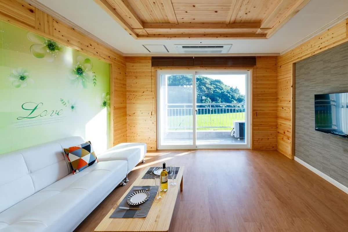 Un salon moderne avec un plafond en bois, un canapé blanc, un téléviseur à écran plat fixé au mur et une porte-fenêtre coulissante donnant sur un espace extérieur.