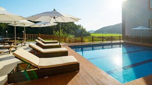 Zona de piscina con sillones de descanso, sombrillas y una terraza de madera.
