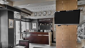 Uma área de recepção com um balcão de madeira, uma televisão fixada na parede e a placa da recepção.