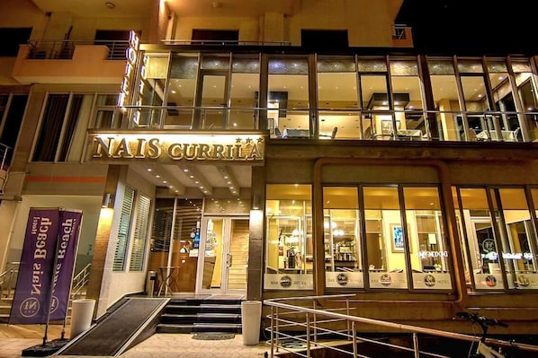 A hotel with a prominent sign reading 'NAIS CURRILA,' featuring a modern facade with large windows and a staircase leading to the entrance.
