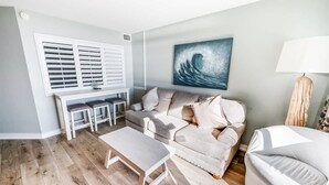 Een woonkamer met een grijze bank, een witte salontafel, een witte fauteuil, een schilderij van een golf en een raam met jaloezieën.