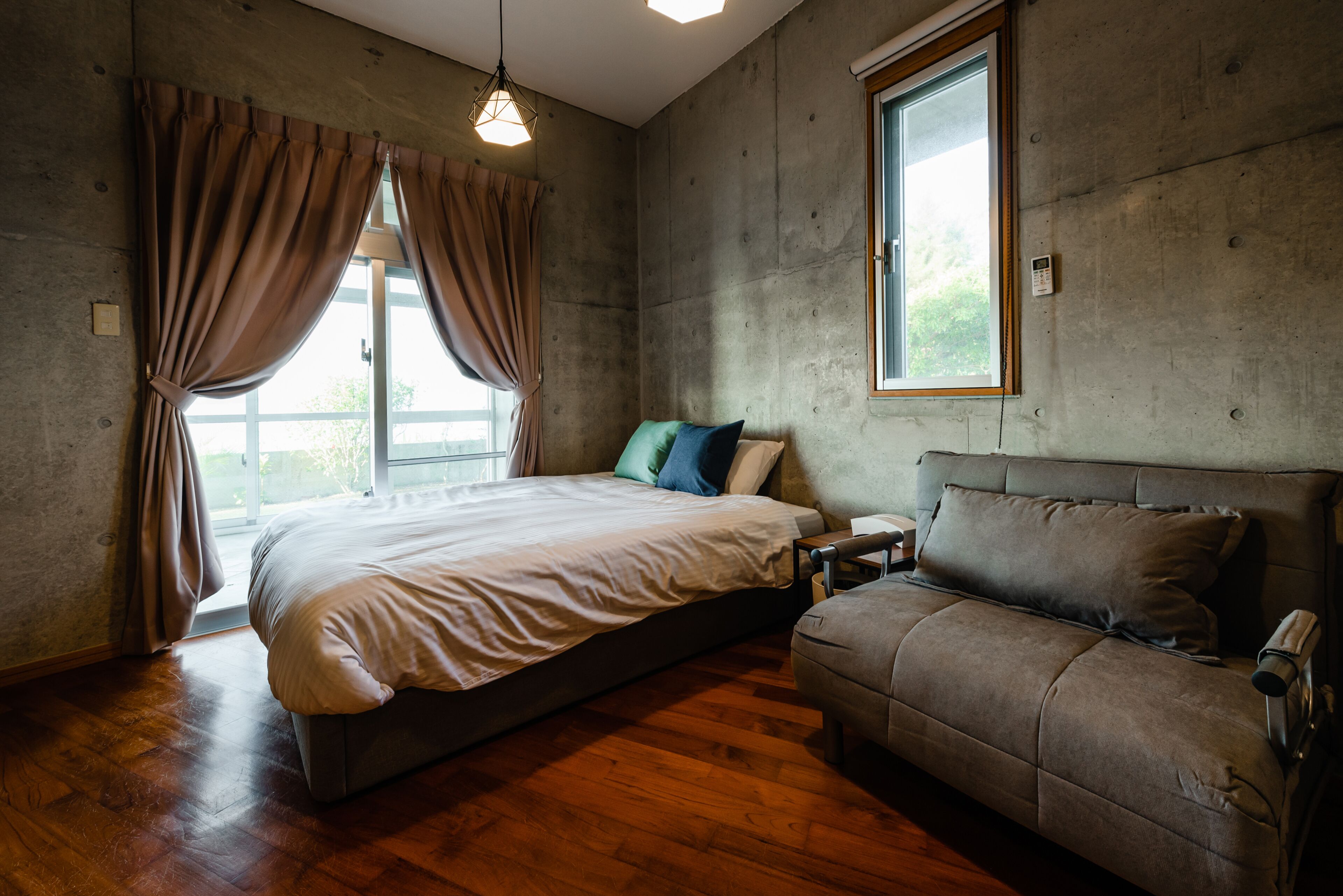 A bedroom with a bed, a sofa, a window, and a concrete wall.