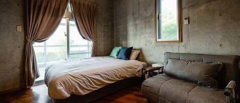 A bedroom with a bed, a sofa, a window, and a concrete wall.