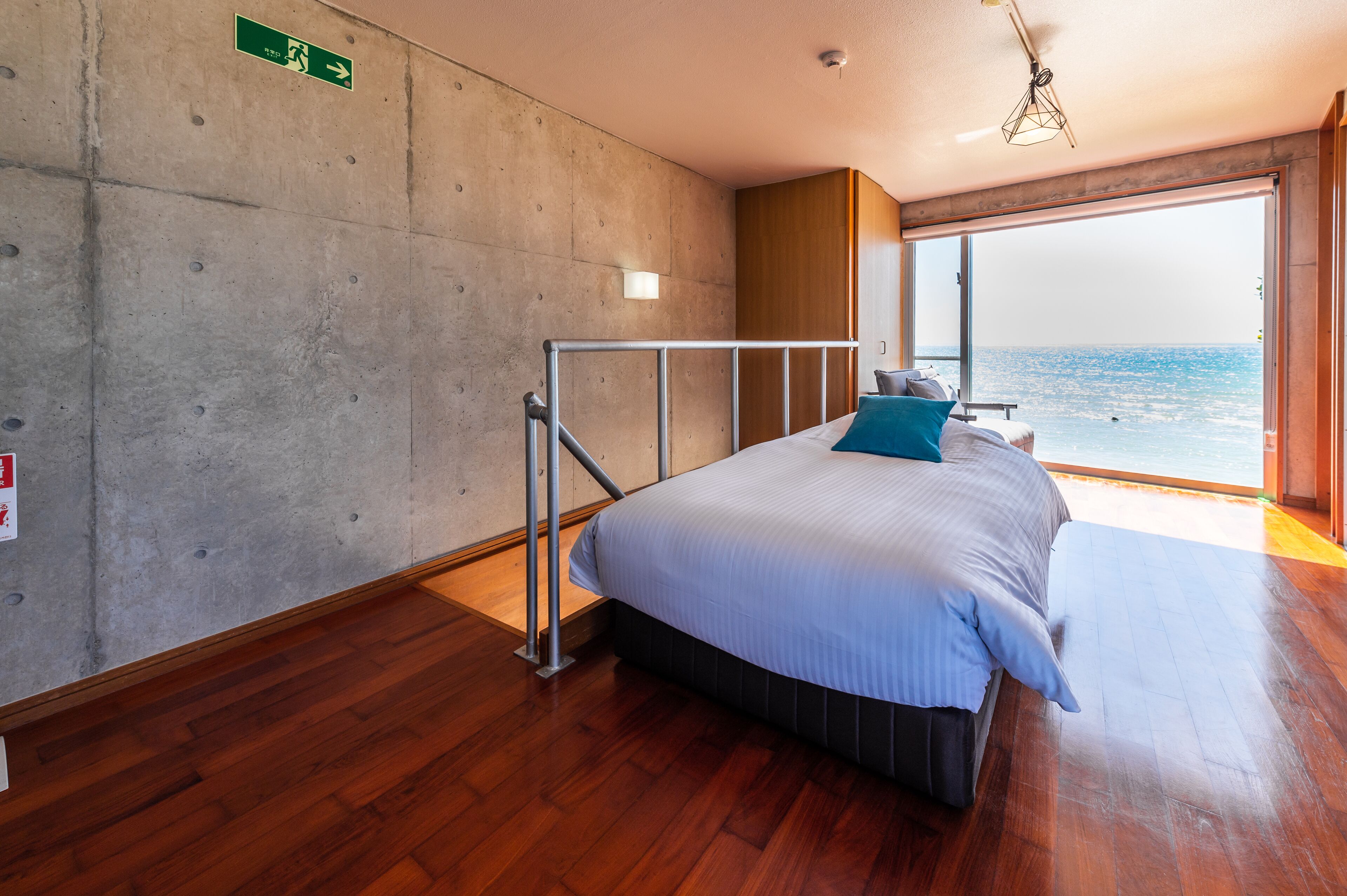 Quarto com cama grande, piso de madeira, parede de concreto e vista para o mar.