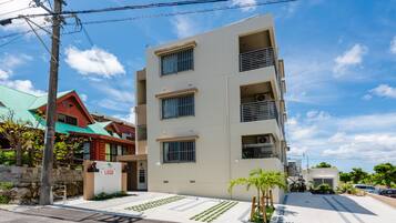 Um edifício com vários andares, telhado plano, entrada visível e uma pequena área de jardim com palmeiras.