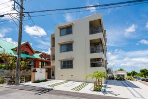 Un edificio de varios pisos con techo plano, entrada visible y un pequeño jardín con palmeras.