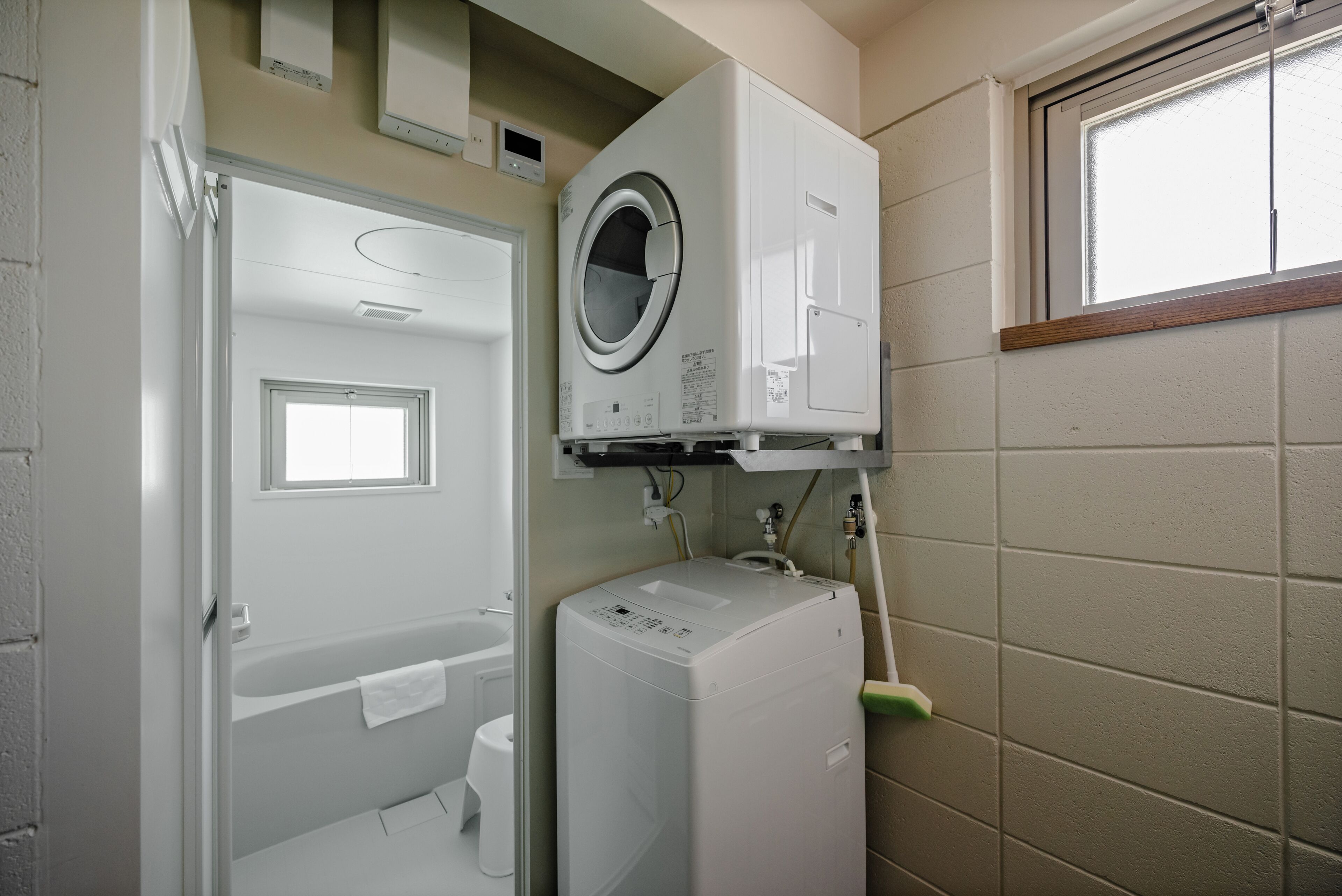 A compact laundry room with a front-loading washer and dryer, a small window, and a door leading to a bathroom.