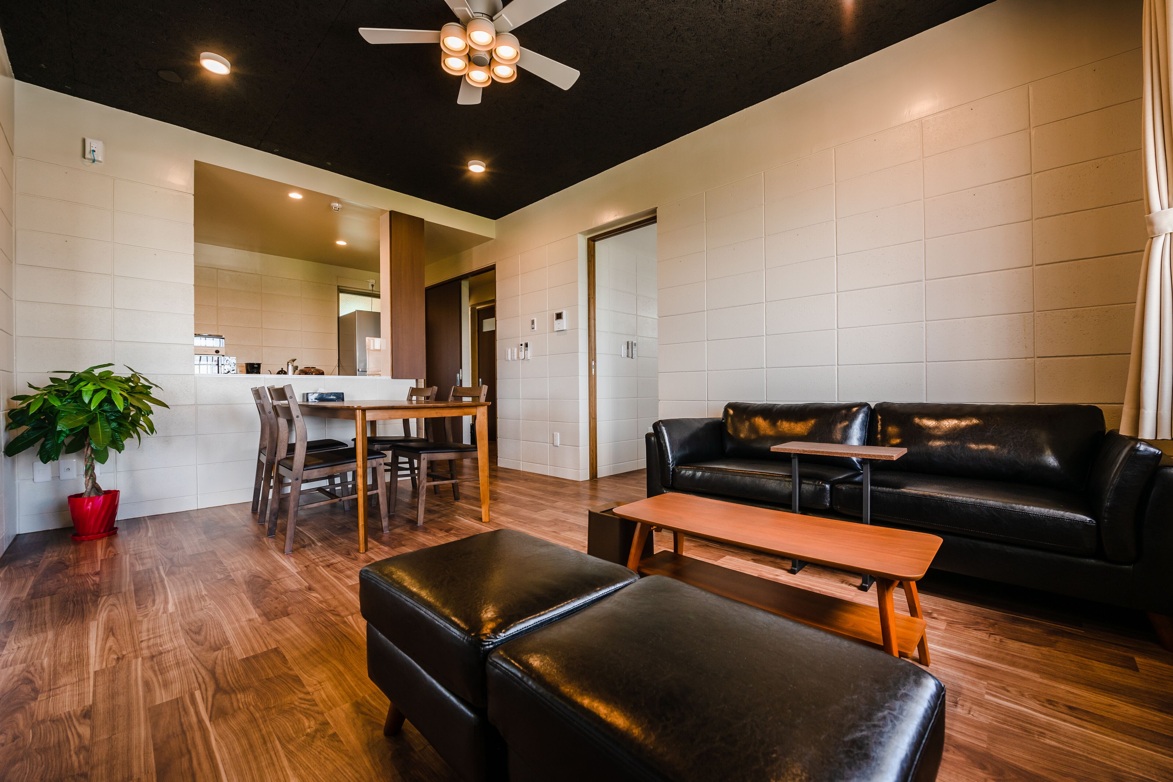 A modern living room with a black leather sofa, wooden coffee table, and ottomans. There is a dining area with a wooden table and chairs, and a potted plant.