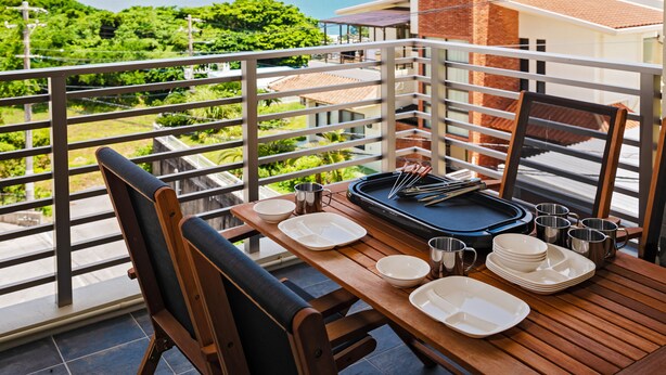 A balcony with a table set for dining, overlooking a scenic view of the sea and greenery. - Lego (Uruma)