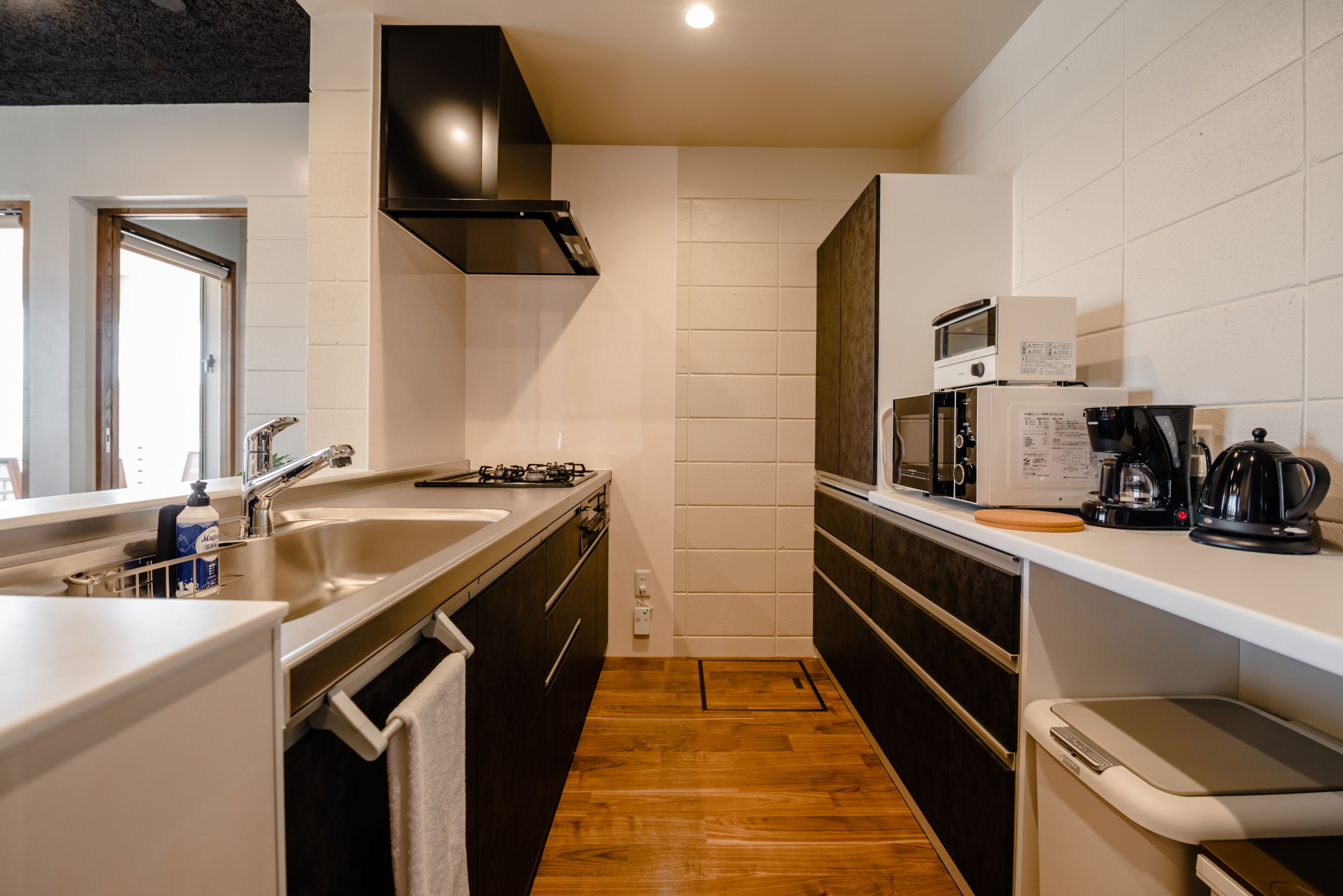 Una cocina moderna con suelo de madera, pileta de acero inoxidable, campana extractora negra, salpicadero de azulejos blancos y una cafetera sobre la encimera.
