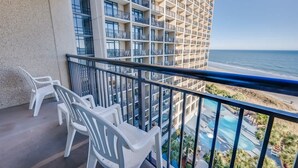 A balcony with white plastic chairs, a view of a building with multiple balconies, and a pool area.