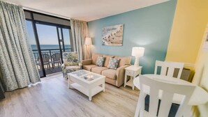 A living room with a sofa, two chairs, a coffee table, and a lamp. There is a large window with curtains and a view of the sea.