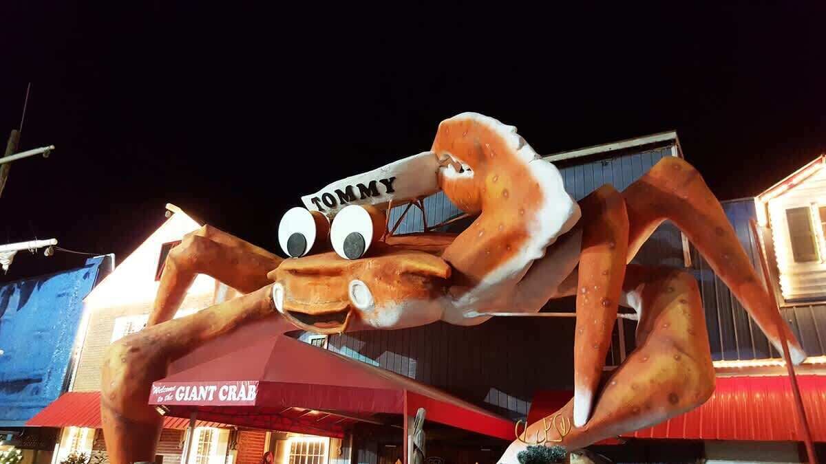 Une grande mascotte représentant un crabe, avec des yeux globuleux et une pancarte sur laquelle on peut lire « TOMMY », devant un bâtiment doté d’un auvent rouge et d’une enseigne indiquant « CRABE GÉANT ».