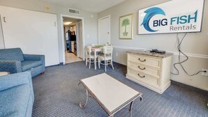 Ett möblerat vardagsrum med en blå soffa, ett litet bord, en vit byrå och en matplats med ett bord och stolar. En skylt för företaget Big Fish Rentals syns på väggen.