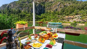 Una mesa de comedor con diversos platos y una tetera en una terraza con vistas a un río, una barca y colinas boscosas al fondo.