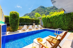 Una piscina con sillas de descanso, una pequeña mesa con un frutero, y montañas al fondo.