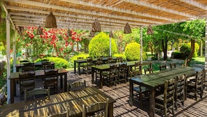 Un patio al aire libre con mesas y sillas de madera, rodeado de abundante follaje y plantas en flor.