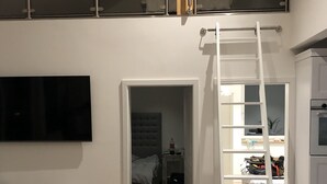A modern apartment with a loft bedroom, a living room with a sofa and a TV, and a kitchen area.