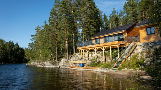 A cabin by a lake with a wooden deck, stairs, and a glass enclosure. - Exclusive and modern holiday home "Lilla älg" right on the lake 365 days a year! (Arvika)