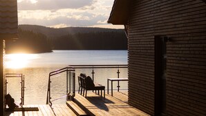 A wooden deck with a railing, a chair, and a table overlooking a lake with a forested hill in the background. - Exclusive and modern holiday home "Lilla älg" right on the lake 365 days a year! (Arvika)