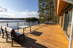 A wooden deck with a bench and table, overlooking a lake with trees in the background. - "Lilla älg," an exclusive and modern vacation home on the lake, available year-round! (Arvika)
