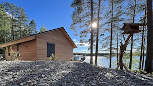 A wooden cabin with a rocky ground, surrounded by tall trees and a lake. - Exclusive and modern holiday home "Lilla älg" right on the lake 365 days a year! (Arvika)