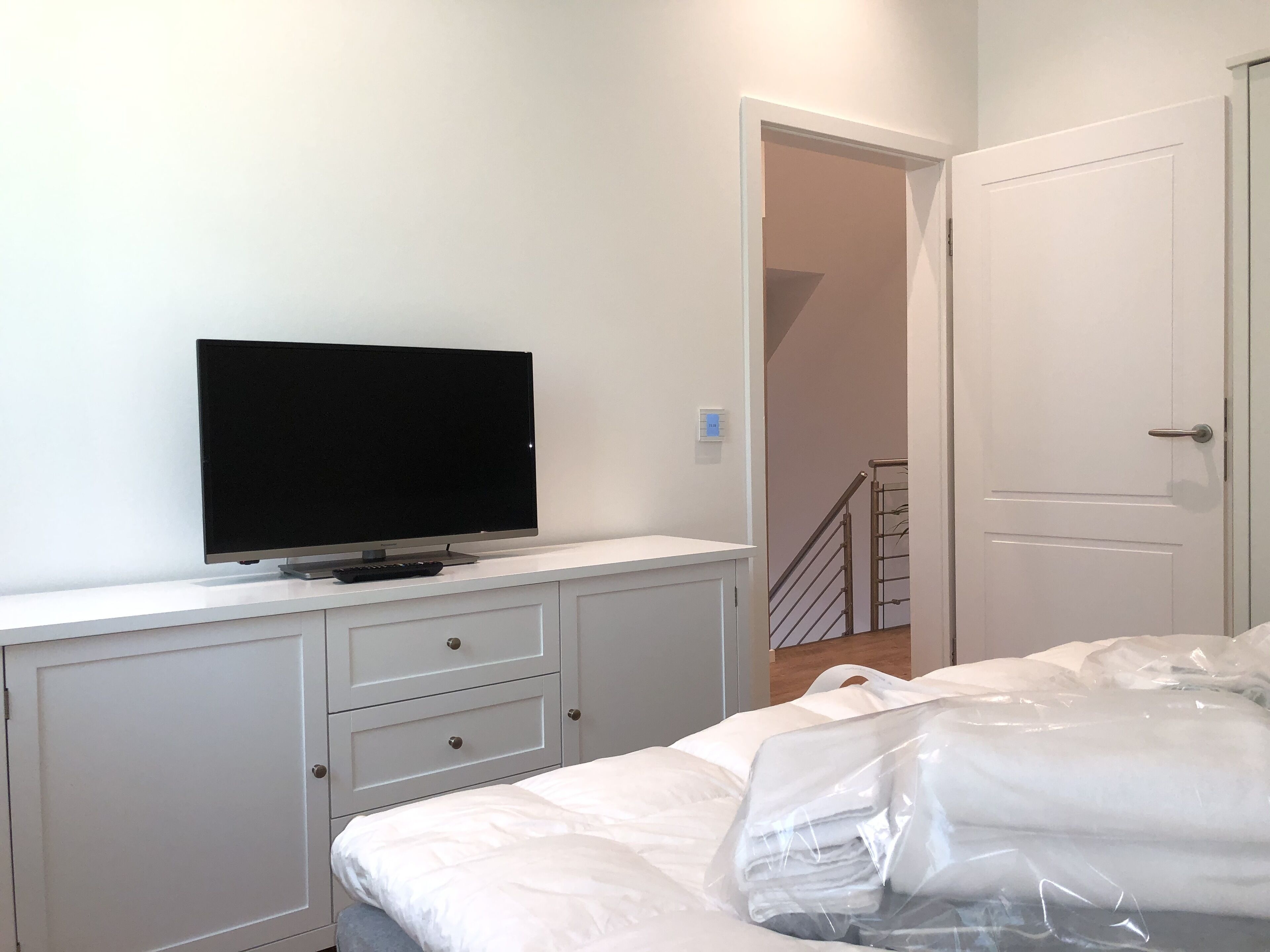 A bedroom with a TV on a white dresser, a bed with white bedding, and a door leading to a staircase.