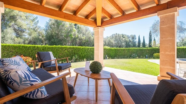 Een terras buiten met houten meubilair, een ronde tafel en uitzicht op een verzorgde tuin.