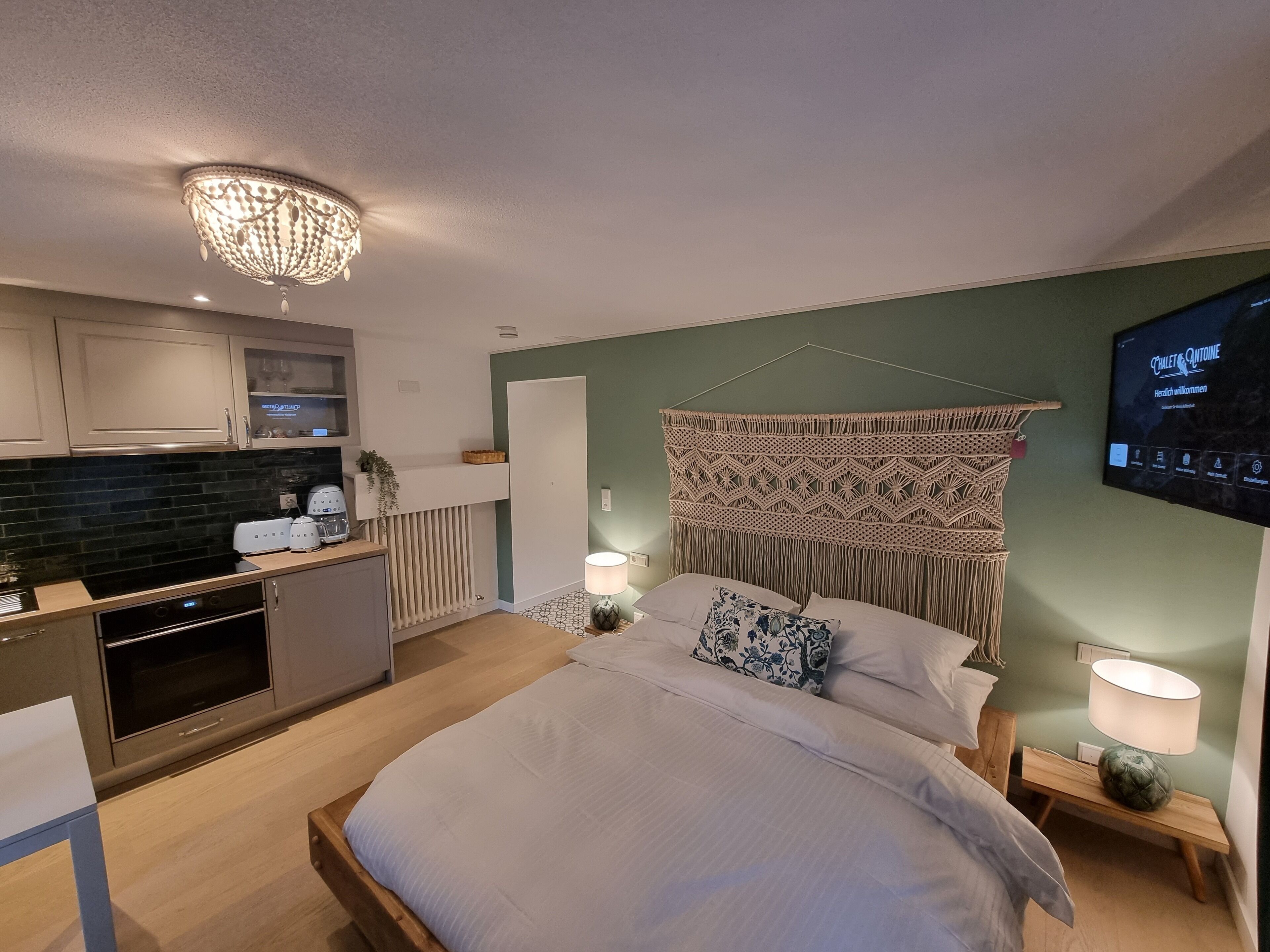 A modern bedroom with a bed, bedside lamps, a TV, and a kitchen area with appliances.