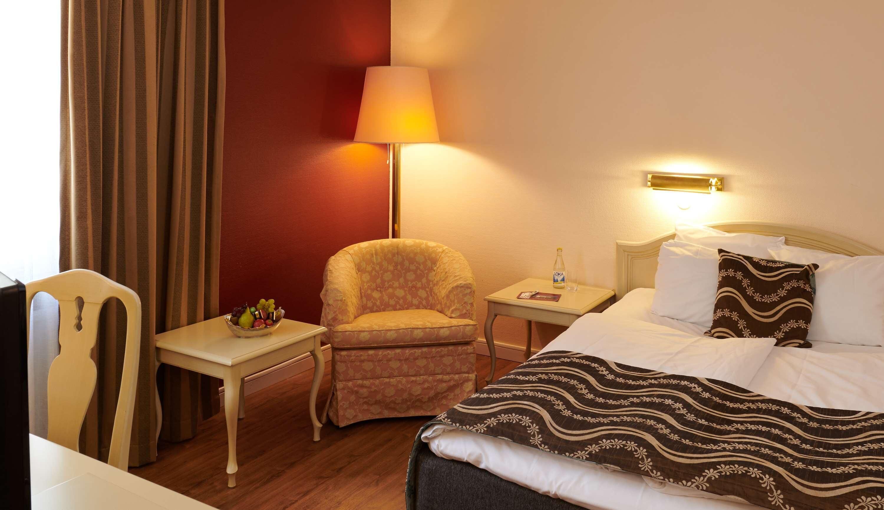 Une chambre d’hôtel avec un lit, une chaise, une petite table avec une lampe et une table de chevet avec une bouteille.