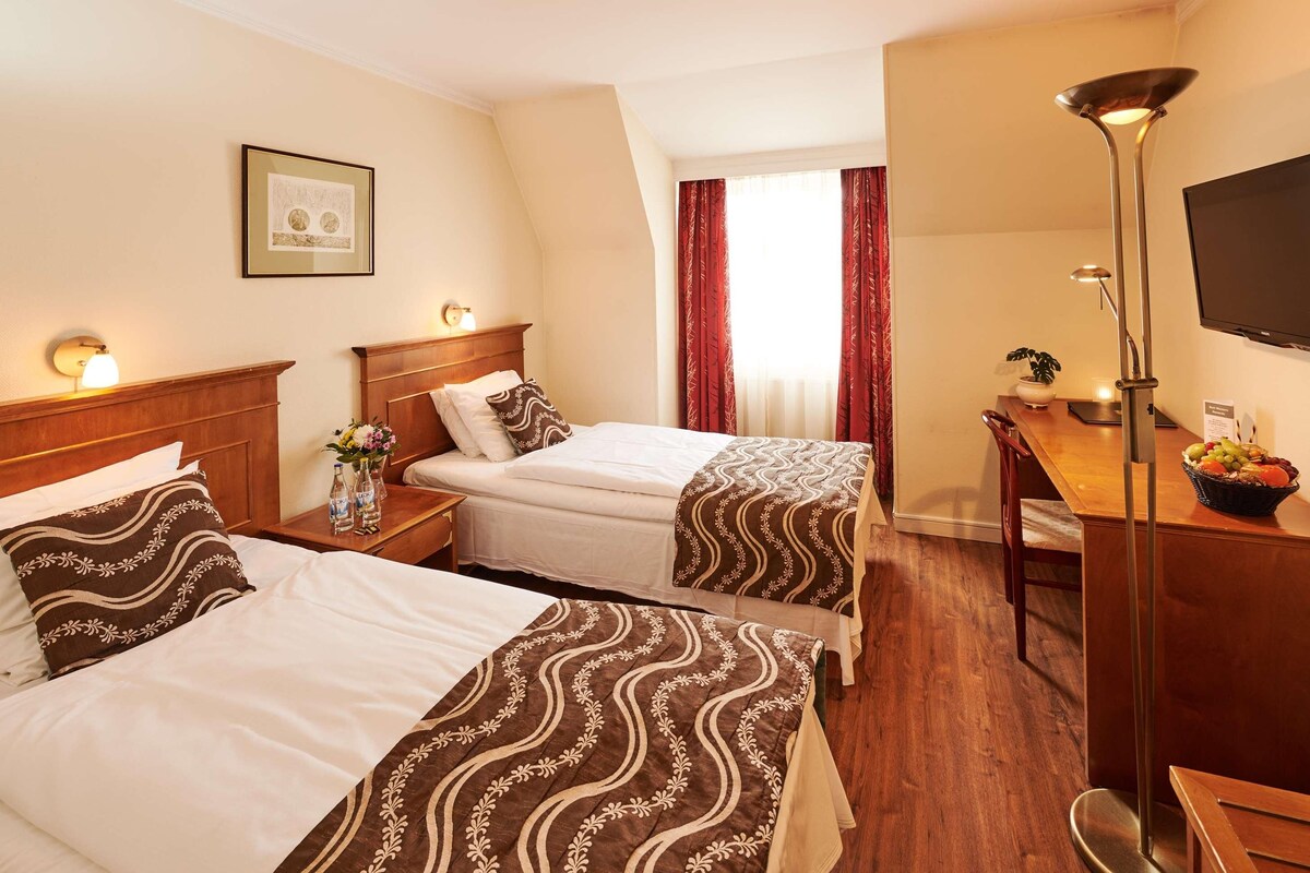 Quarto de hotel com duas camas, uma escrivaninha, televisão e um quadro na parede.