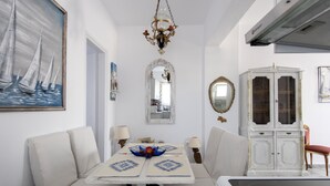 Un coin repas avec une table blanche, des chaises et un lustre. On y trouve un buffet avec des portes en verre et un miroir accroché au mur.