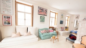 A spacious living room with a white sofa, a teal armchair, and a wooden coffee table. There are large windows providing natural light, and the walls are adorned with various framed artworks.