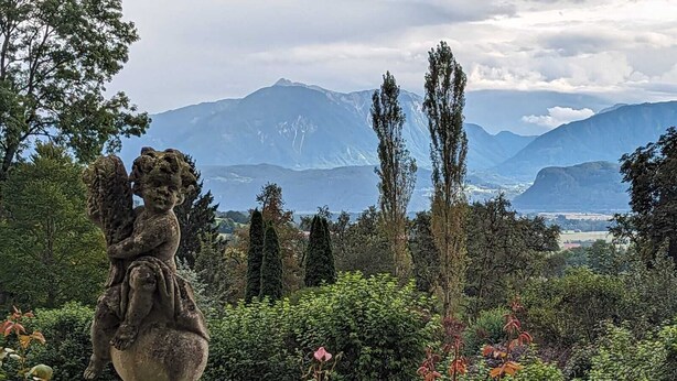 A garden with a statue of a cherub, surrounded by lush greenery and flowering plants, with a mountainous landscape in the background.
