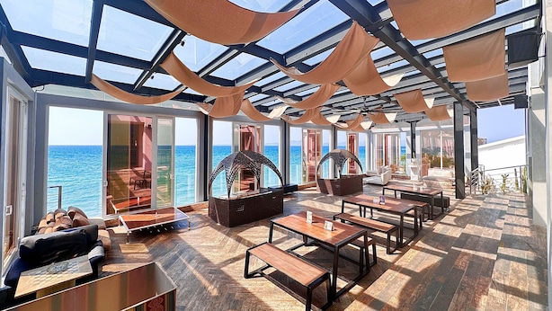 Een zonnig buitenterrein met tafels en stoelen, grote ramen met uitzicht op de oceaan en een glazen dakconstructie.