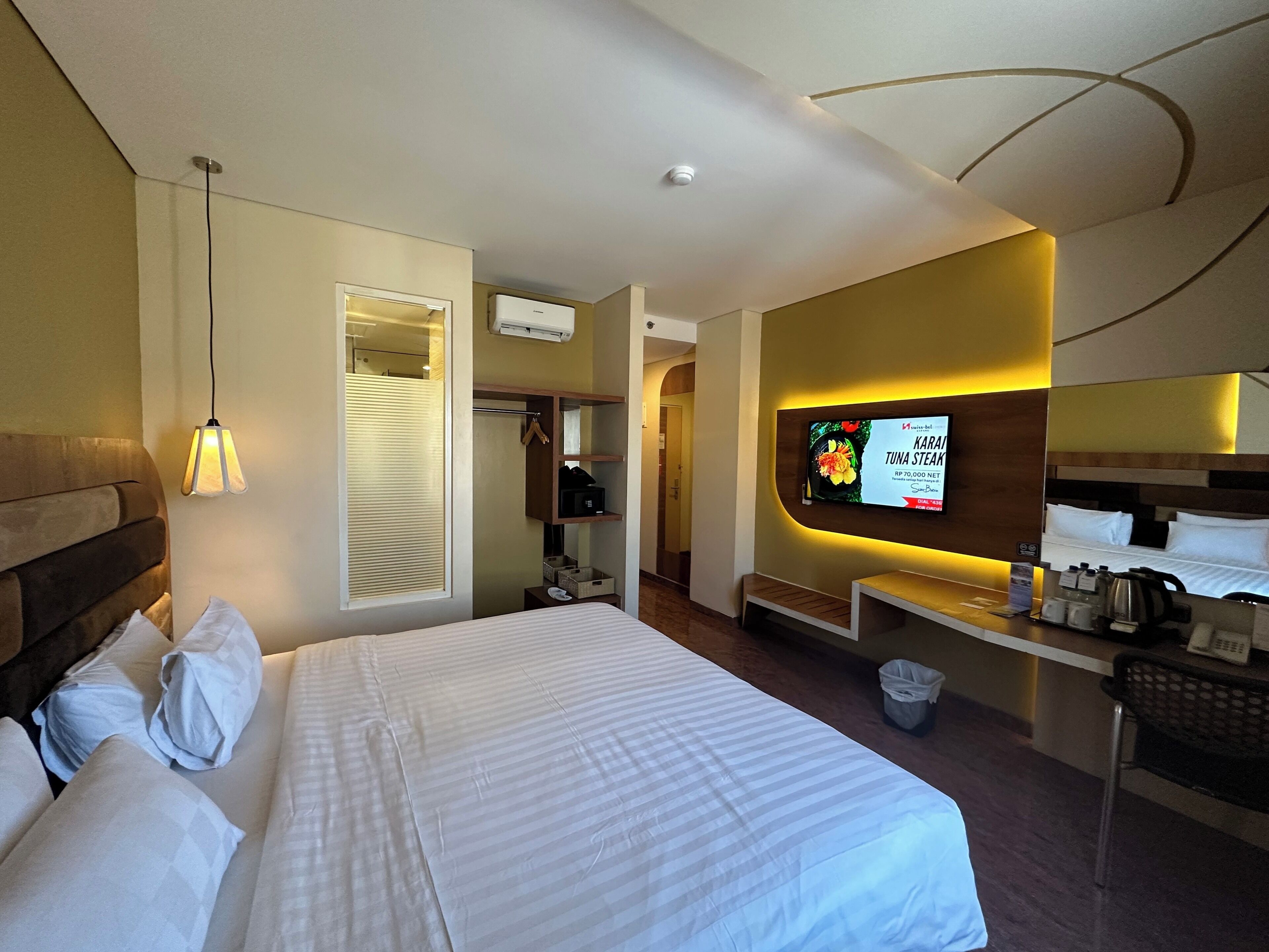 A hotel room with a large bed, a desk with a coffee maker, a TV displaying a menu, and a mirror above the bed.