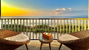 Een balkon met rieten stoelen en een kleine tafel, met uitzicht op een strand en een helder uitzicht over de zee en verre bergen.