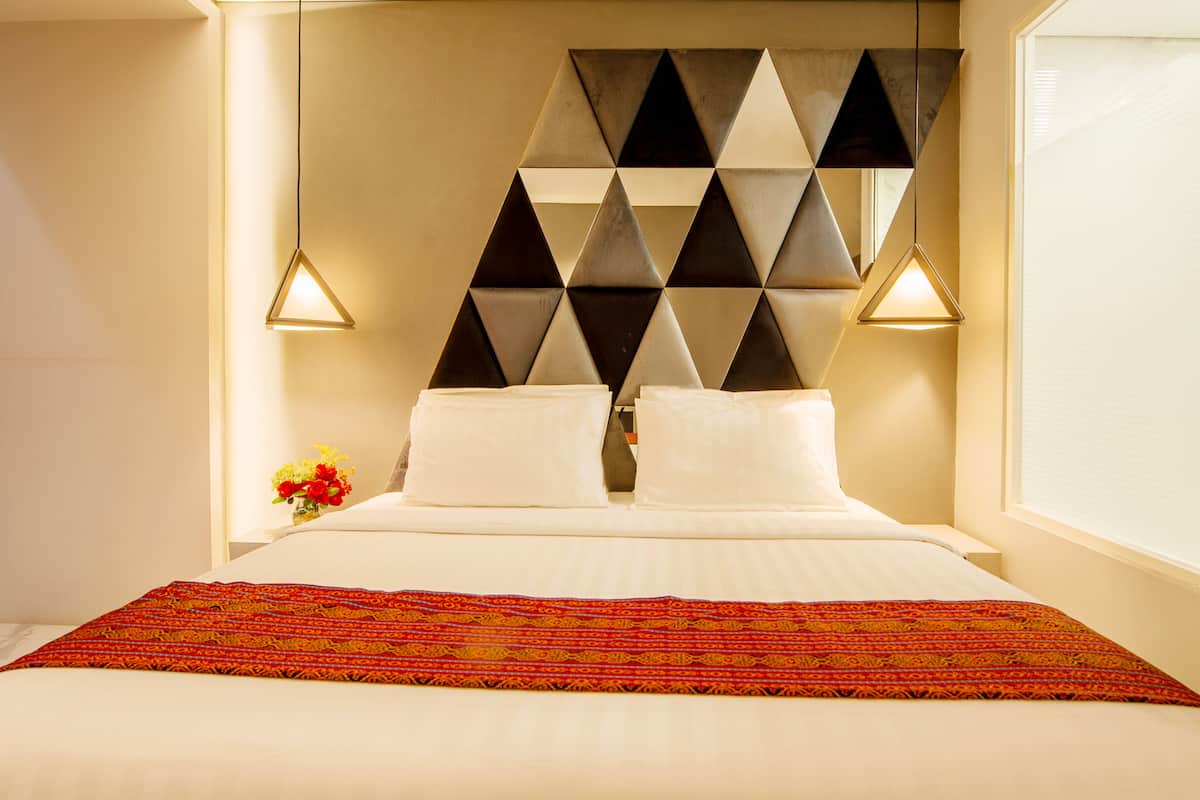 Une chambre d’hôtel avec une tête de lit à motifs géométriques, deux lampes de chevet, un lit soigneusement fait avec du linge de lit blanc et un plaid coloré.