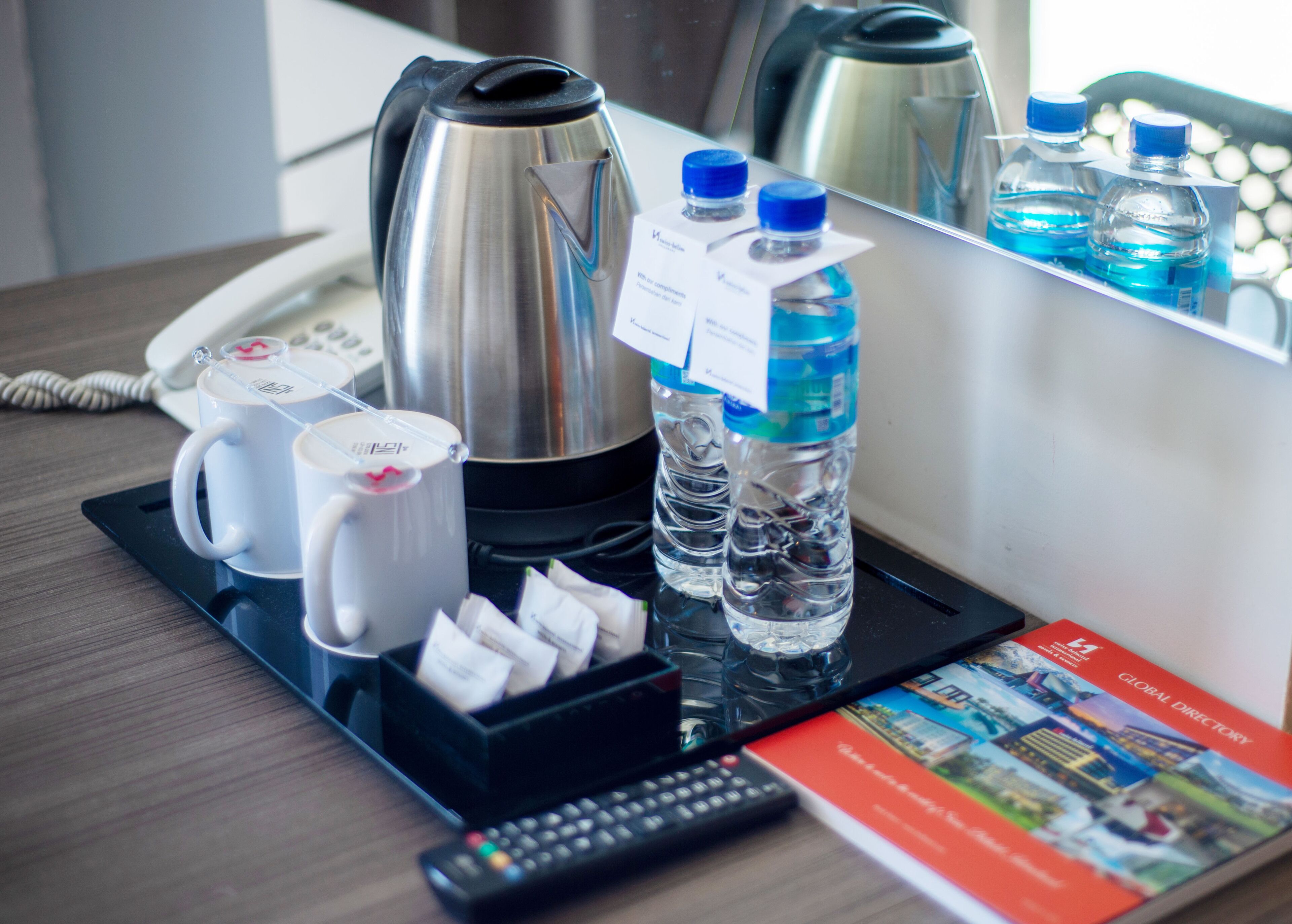 A hotel room with a coffee maker, two mugs, a tray with tea bags, two water bottles, a remote control, and a Global Directory book.