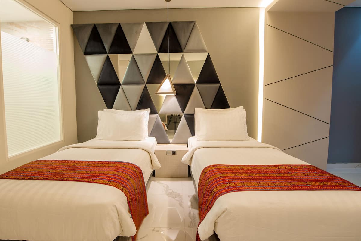 Une chambre d’hôtel avec deux lits, une tête de lit à motifs géométriques, une lampe de chevet et une fenêtre avec des stores.