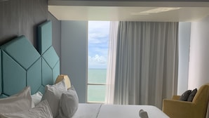 Een hotelkamer met een groot bed, een hoofdbord, uitzicht op zee en een gele fauteuil.