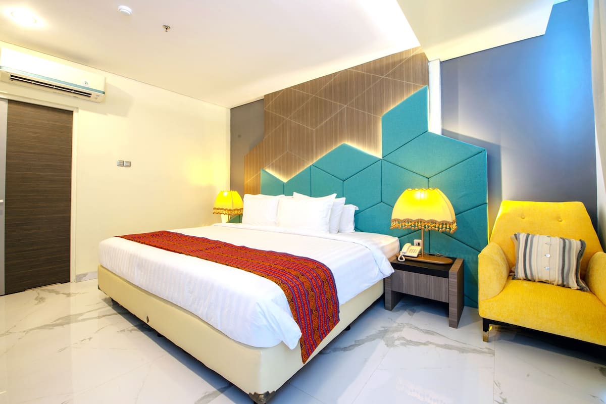 Une chambre d’hôtel moderne avec un grand lit, une table de chevet avec une lampe, un fauteuil jaune et un motif géométrique sur le mur.