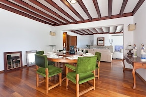 Een eetruimte met een houten tafel en groen gestoffeerde stoelen, een keuken op de achtergrond en houten balken aan het plafond.