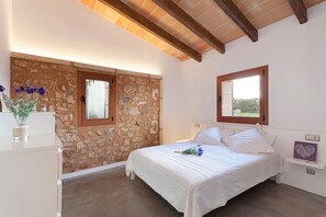 Une chambre avec un mur en pierre, un lit avec une literie blanche, une fenêtre et une petite étagère ornée d’un objet décoratif en forme de cœur.